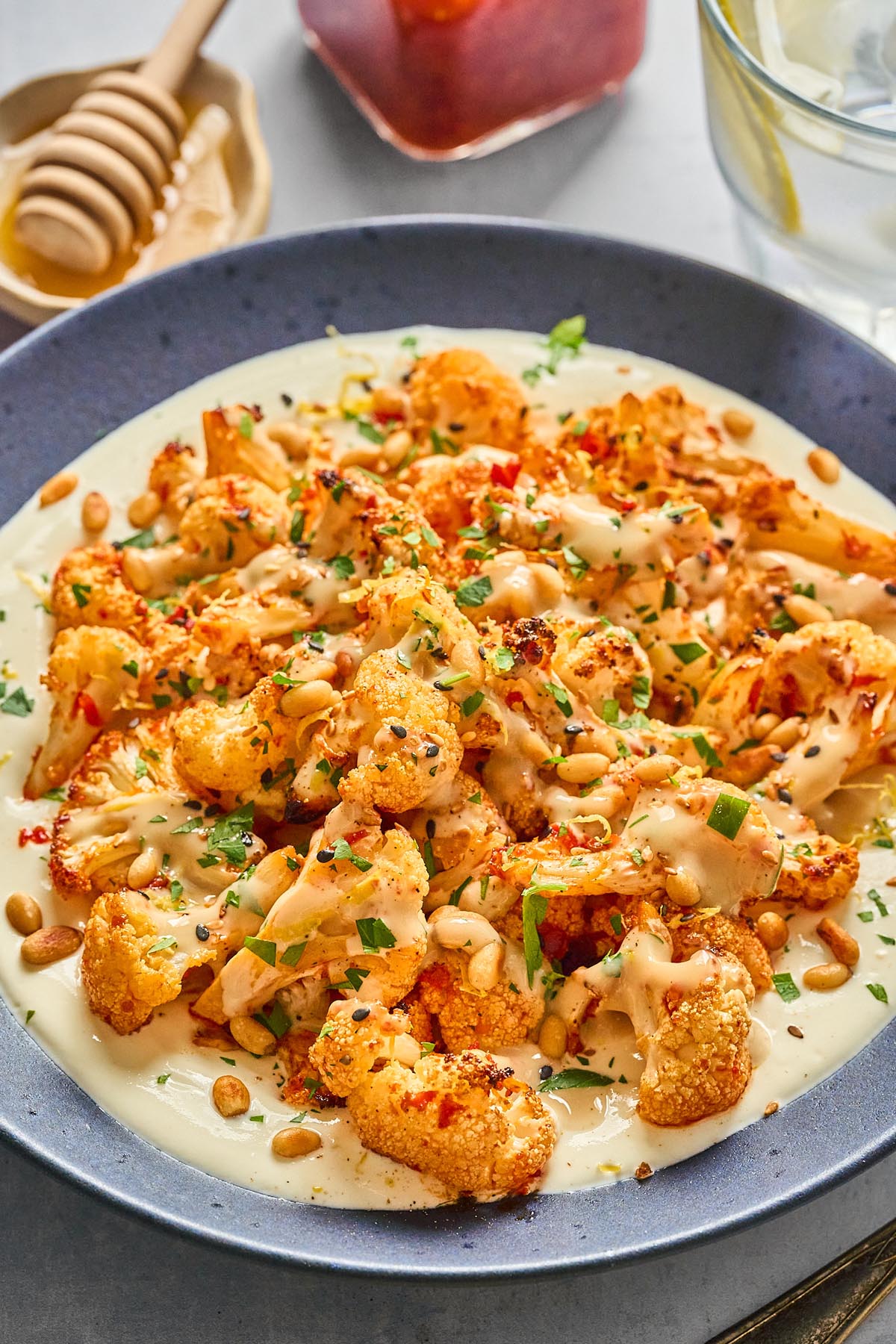 Close-up of harissa roasted cauliflower served over whipped cottage cheese, drizzled with honey-tahini sauce, garnished with pine nuts, parsley, and sesame seeds.