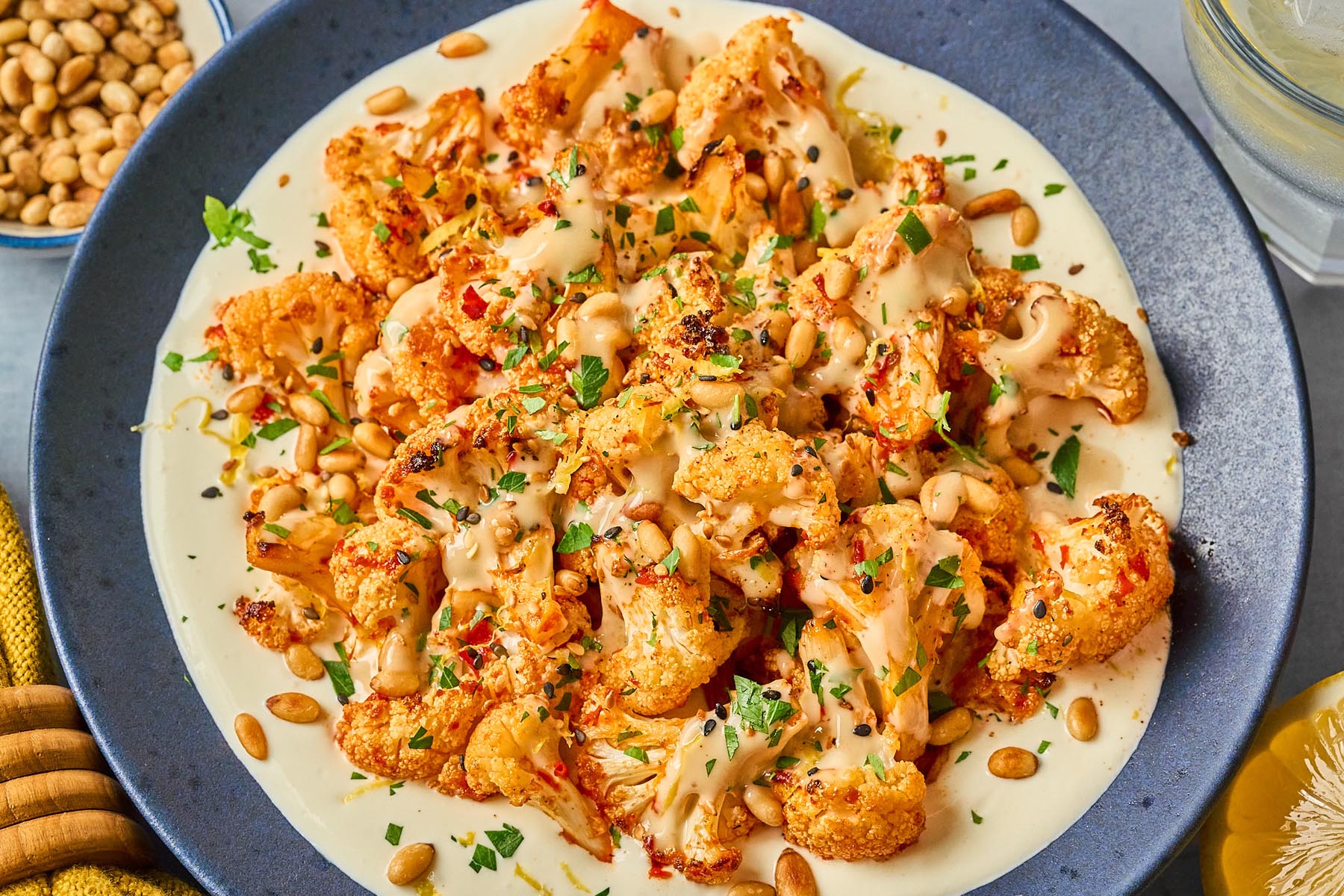 Harissa roasted cauliflower served over whipped cottage cheese, drizzled with honey-tahini sauce and topped with toasted pine nuts, parsley, and sesame seeds on a dark plate.