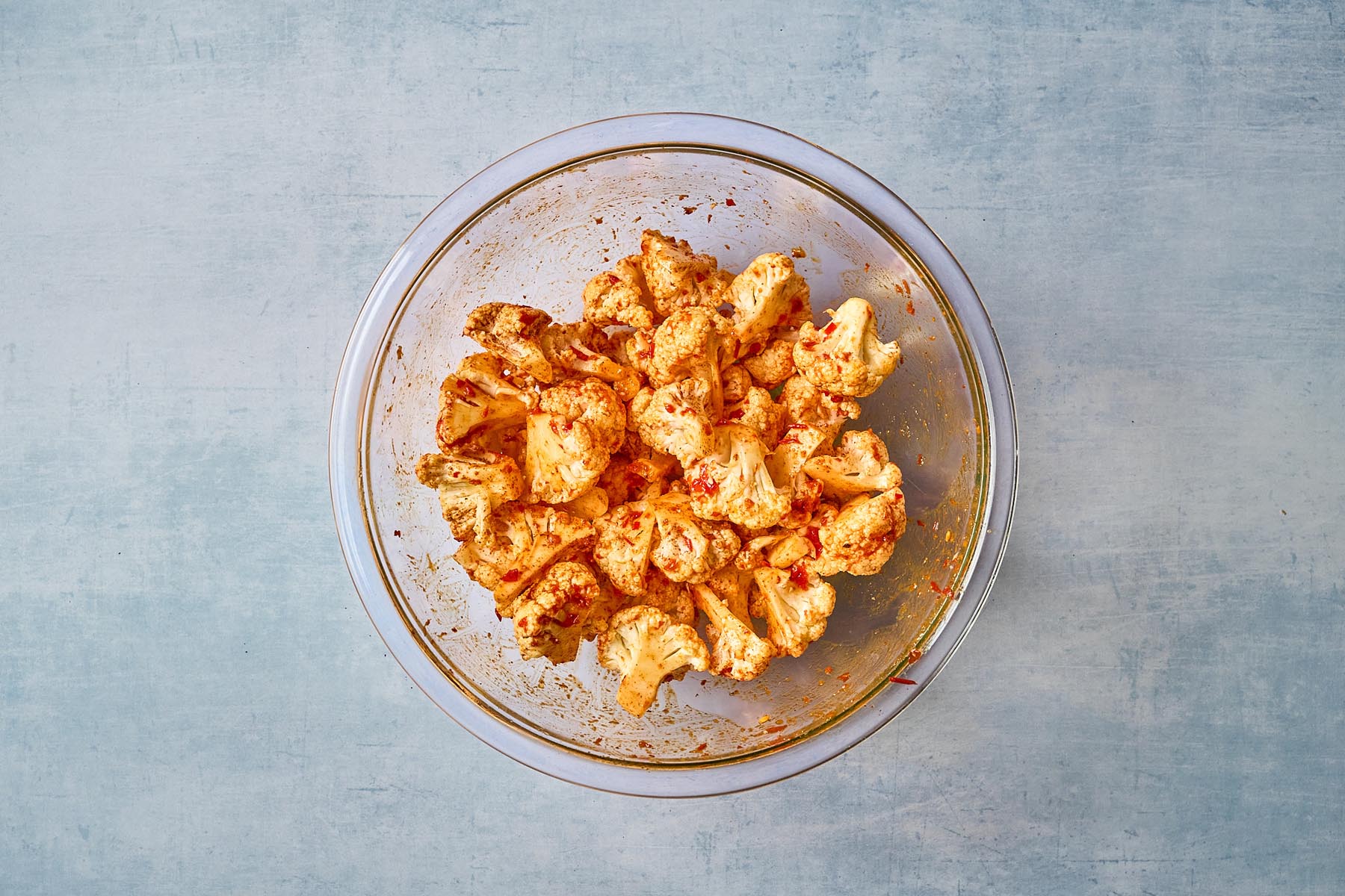 Cauliflower florets tossed with harissa paste, olive oil, and seasoning in a glass bowl, coated evenly and ready for roasting.