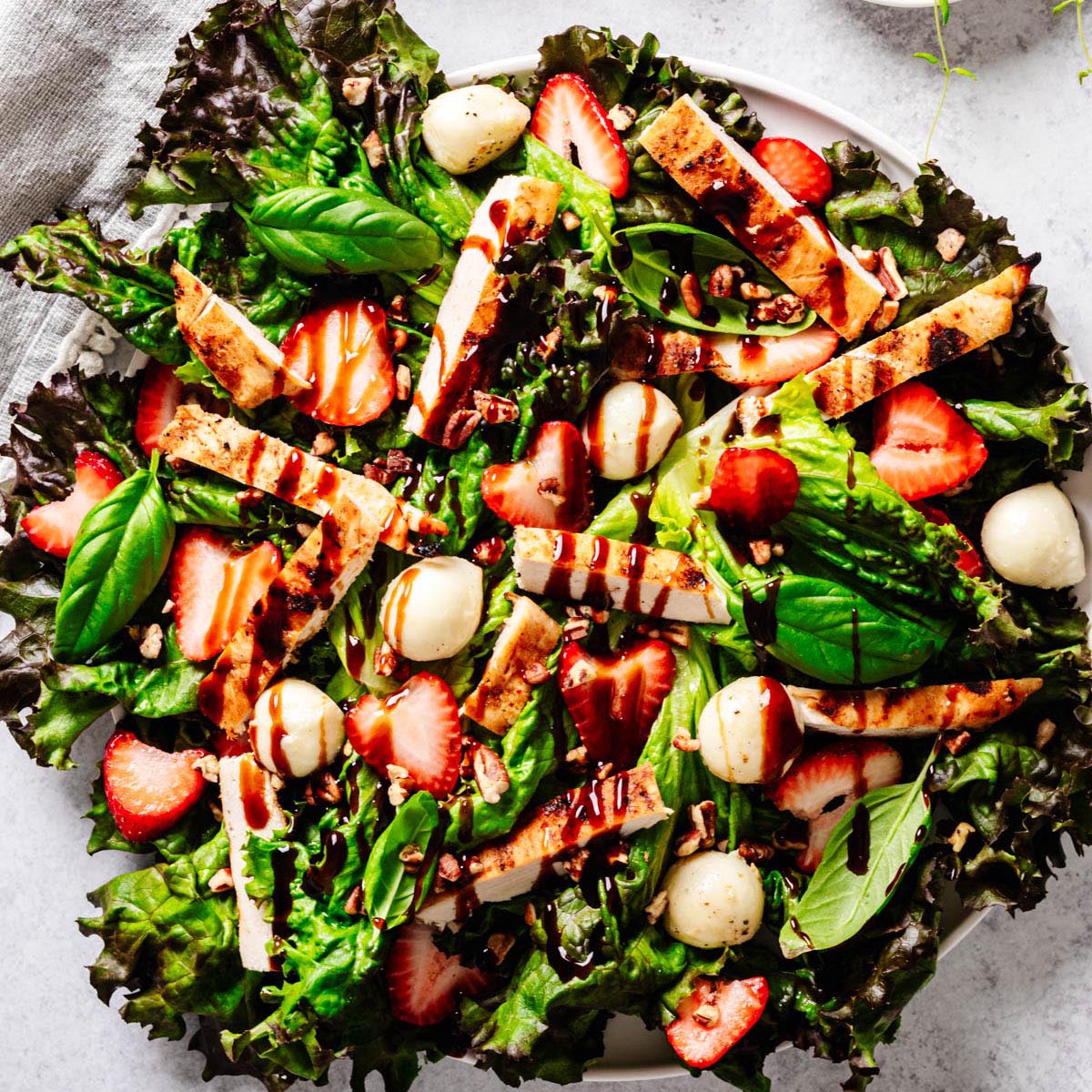 Strawberry chicken Caprese salad served on a white plate, featuring mixed greens, grilled chicken strips, fresh strawberries, mozzarella balls, chopped pecans, basil leaves, and a drizzle of balsamic glaze.