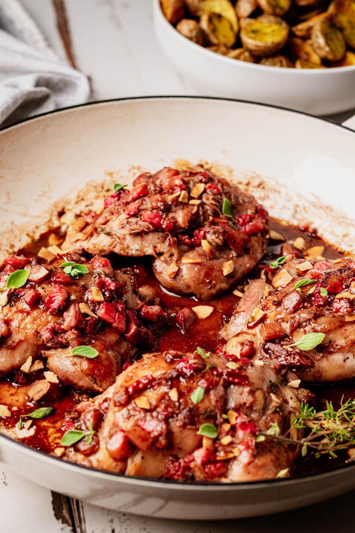 Close-up of strawberry chicken in a white skillet, glazed with strawberry balsamic sauce and topped with toasted almonds and herbs, with a bowl of roasted potatoes in the background.