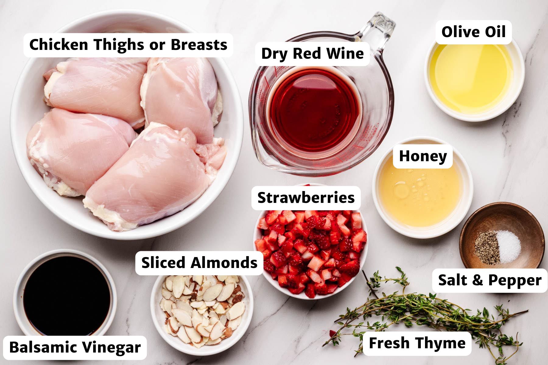 Labeled ingredients for strawberry chicken arranged on a white surface, including chicken thighs or breasts, dry red wine, olive oil, honey, strawberries, balsamic vinegar, sliced almonds, fresh thyme, salt, and pepper.