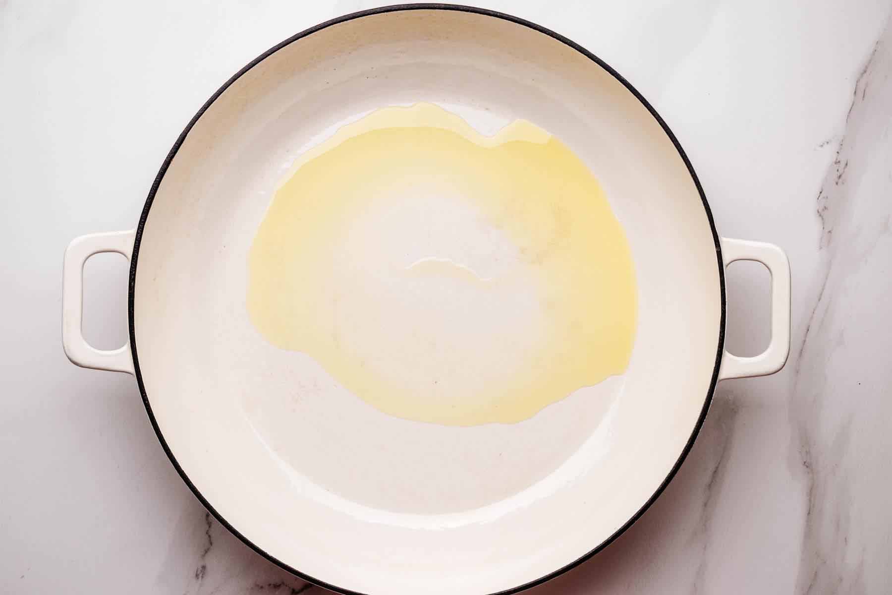 Olive oil heating in a large white skillet on a marble surface.