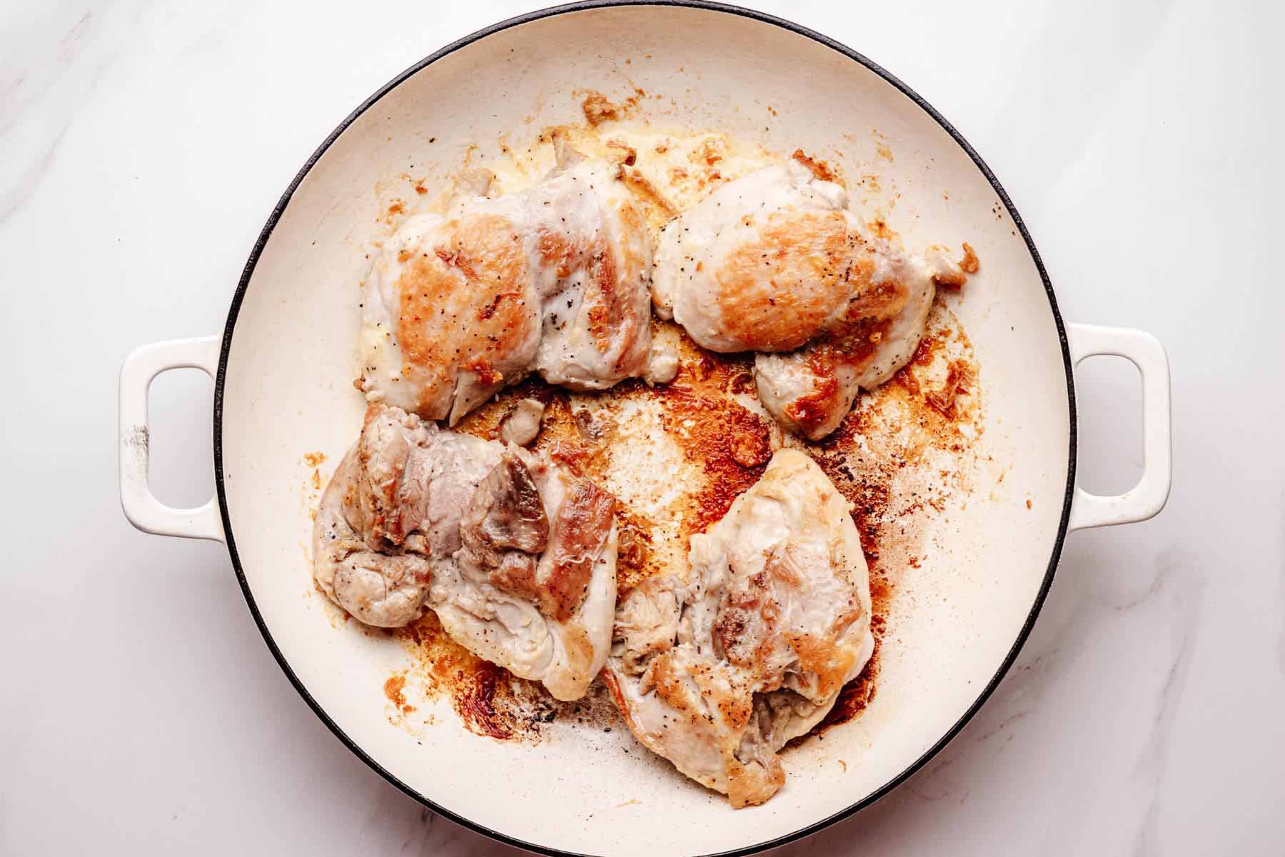 Chicken thighs searing in a white skillet until golden brown, with browned bits forming on the bottom of the pan.