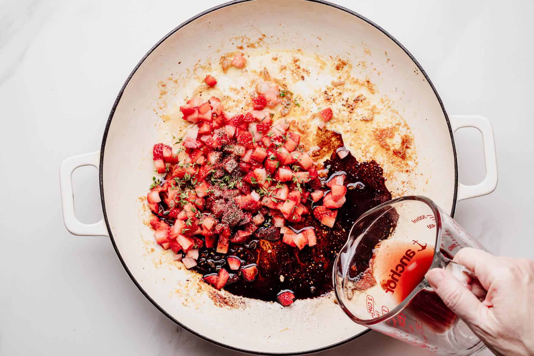 Diced strawberries, thyme, and balsamic vinegar in a skillet with red wine being poured in to begin making the glaze.