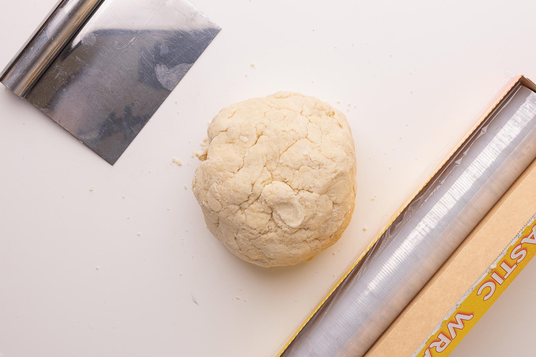 Ball of pie dough on a white surface with a bench scraper and roll of plastic wrap beside it, ready to divide and chill.