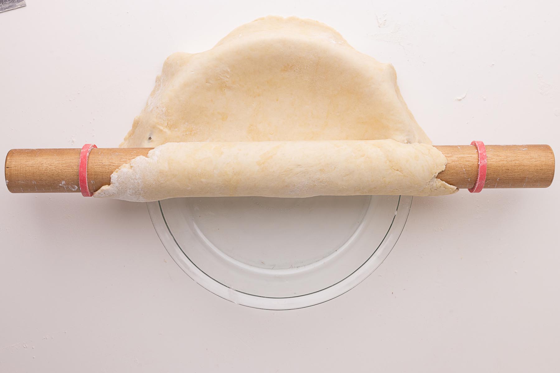 Rolled-out pie dough being unrolled from a wooden rolling pin into a glass pie plate.