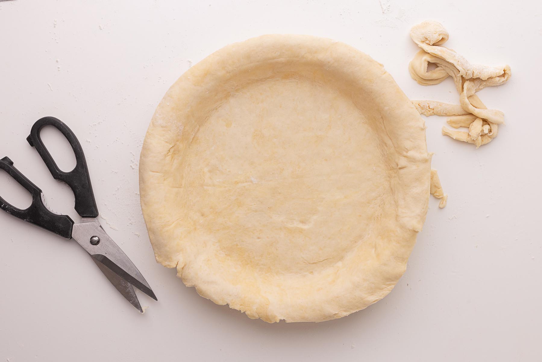 "Unbaked pie crust trimmed to fit a glass pie dish, with kitchen shears and dough scraps on a white surface.