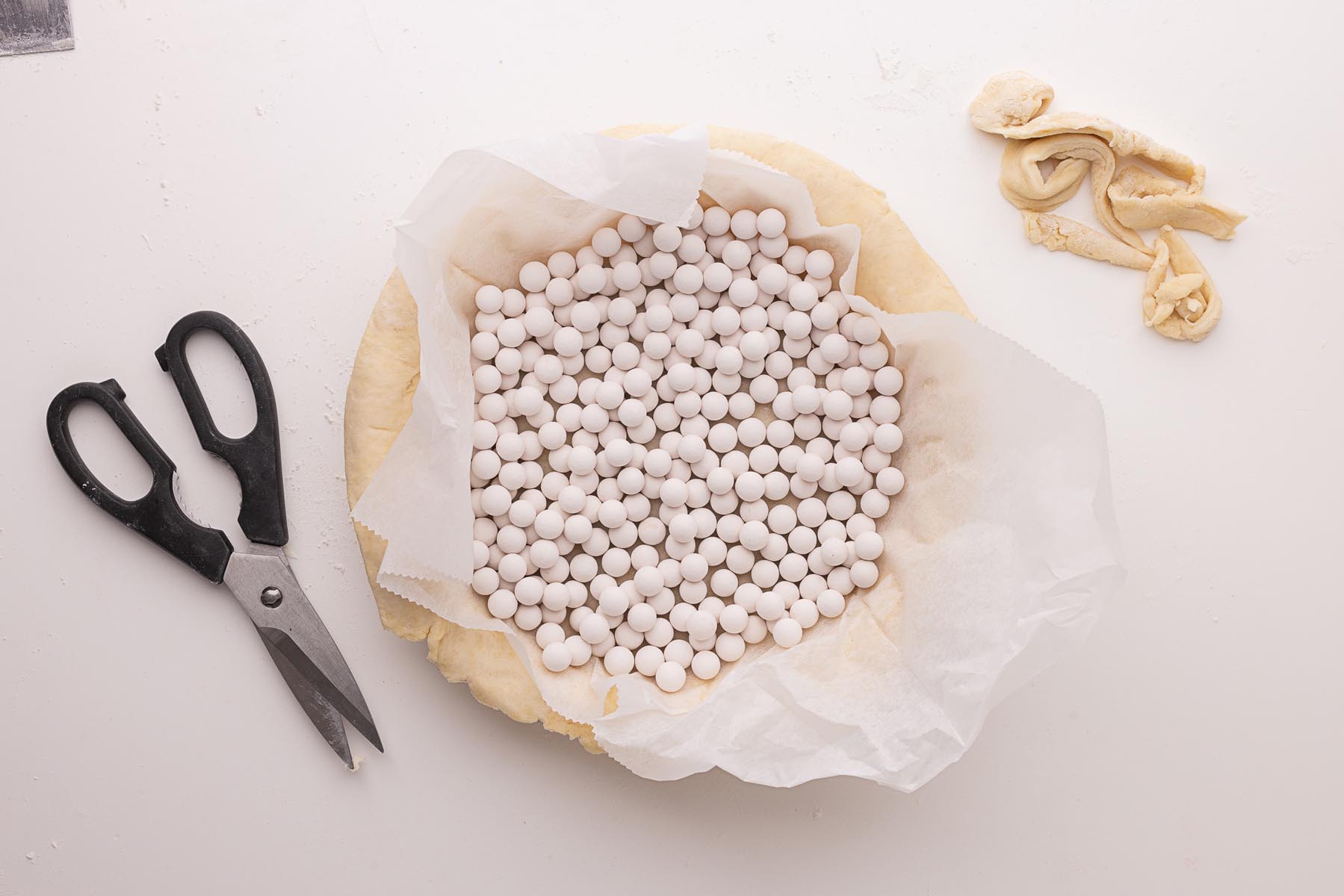 Unbaked pie crust lined with parchment paper and filled with ceramic pie weights, surrounded by dough scraps and kitchen shears on a white surface.