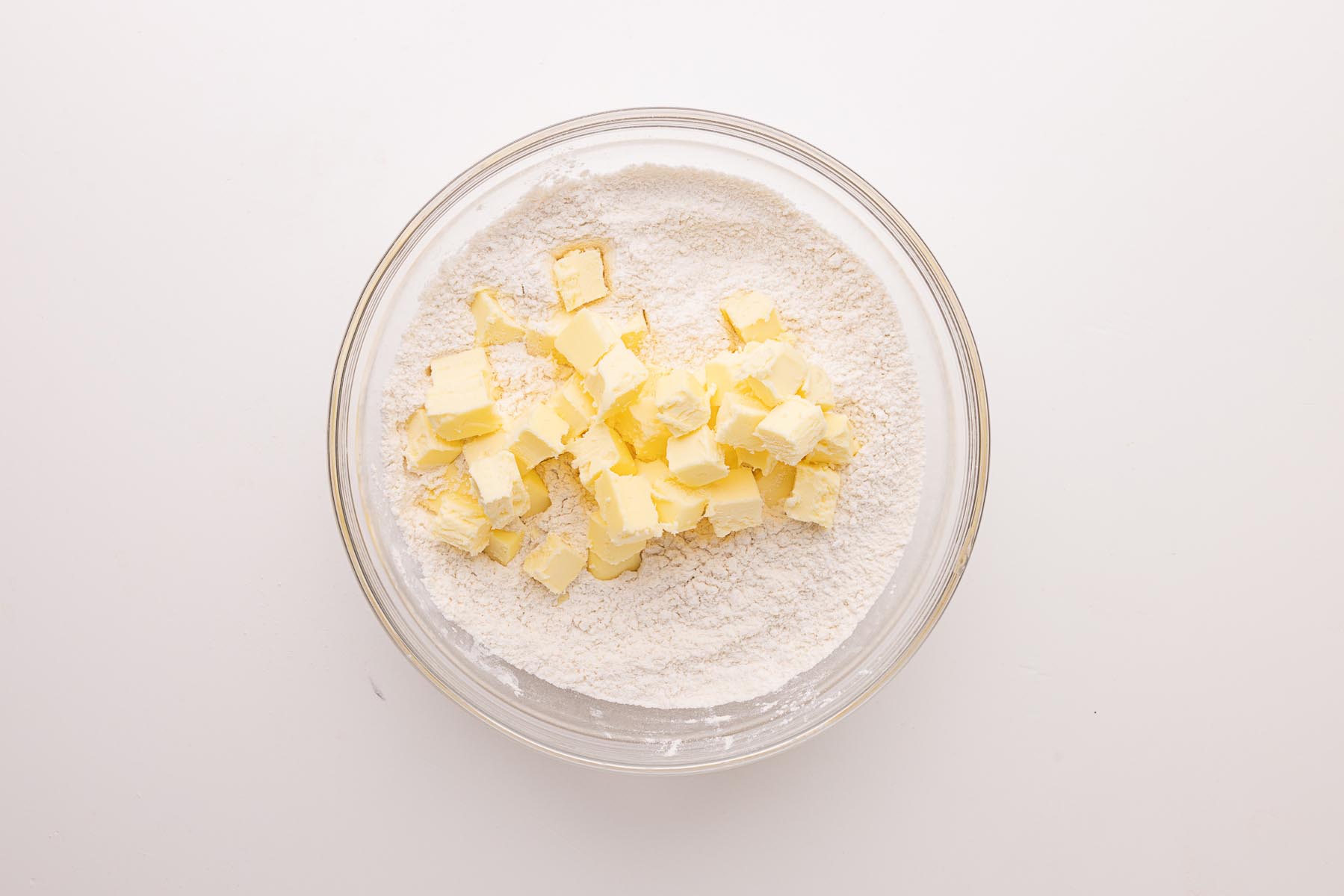 Cold cubes of butter added to a glass bowl of flour mixture, ready to be pressed and flaked by hand for pie crust.