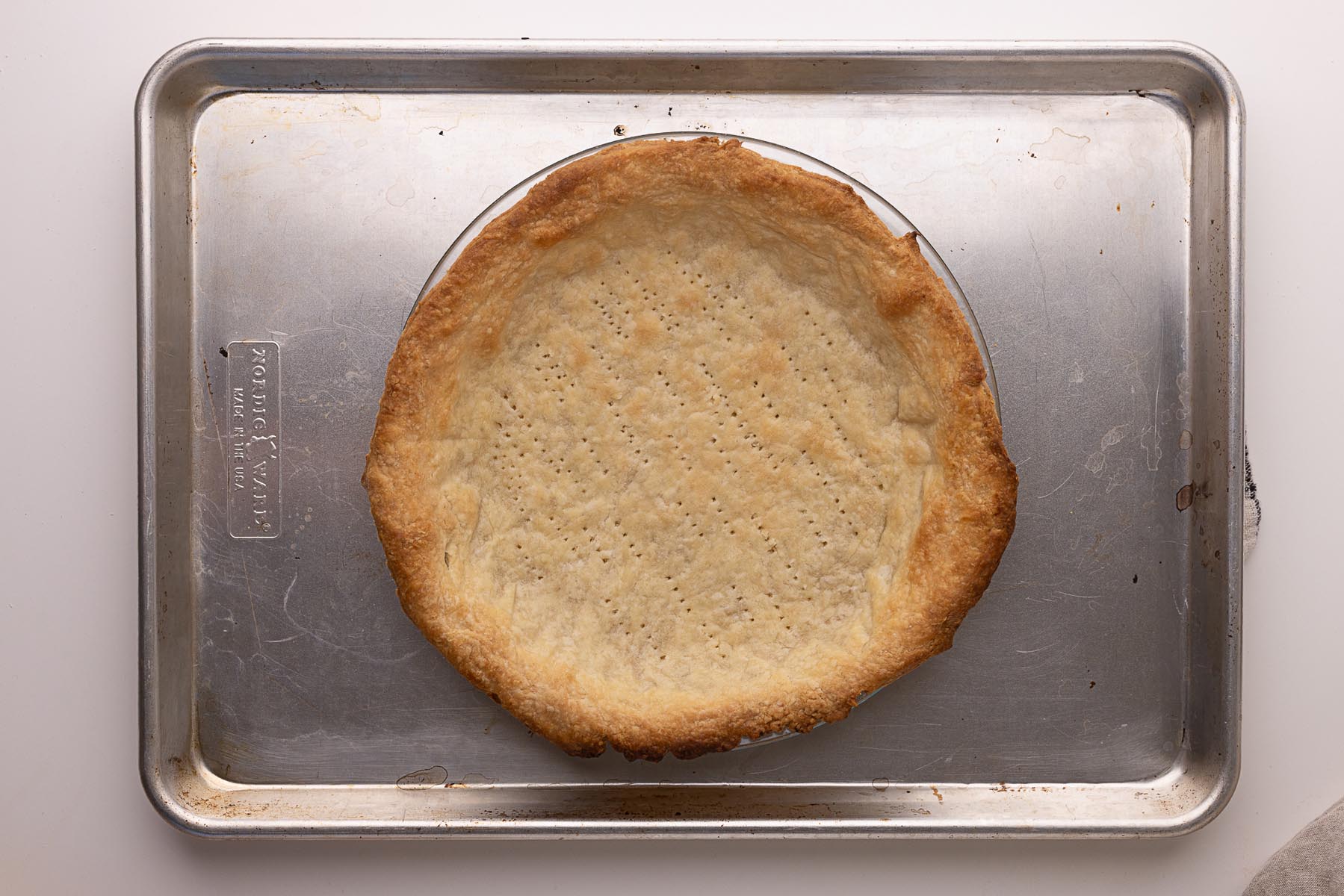Fully baked pie crust in a glass pie dish, resting on a metal baking sheet. The crust is golden brown with fork marks across the bottom.