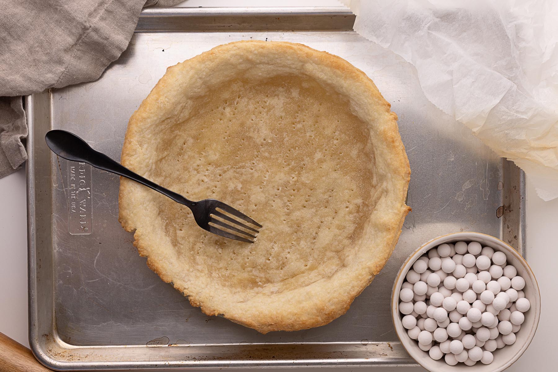Partially baked pie crust in a metal pie dish on a baking sheet, with a fork resting inside the crust. A small bowl of ceramic pie weights and used parchment paper sit nearby.