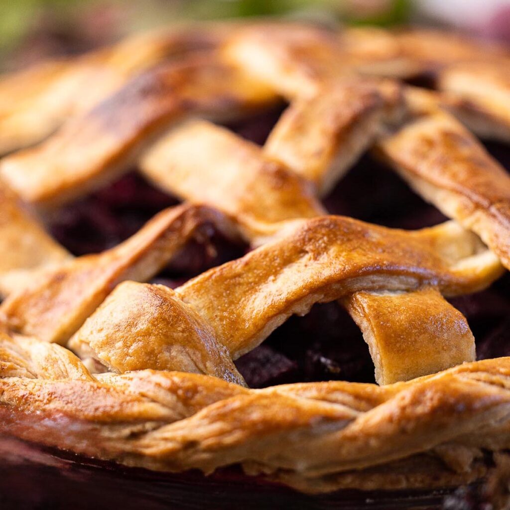 Golden, flaky lattice pie crust shown in close-up, revealing crisp texture and deep purple fruit filling beneath the woven top.