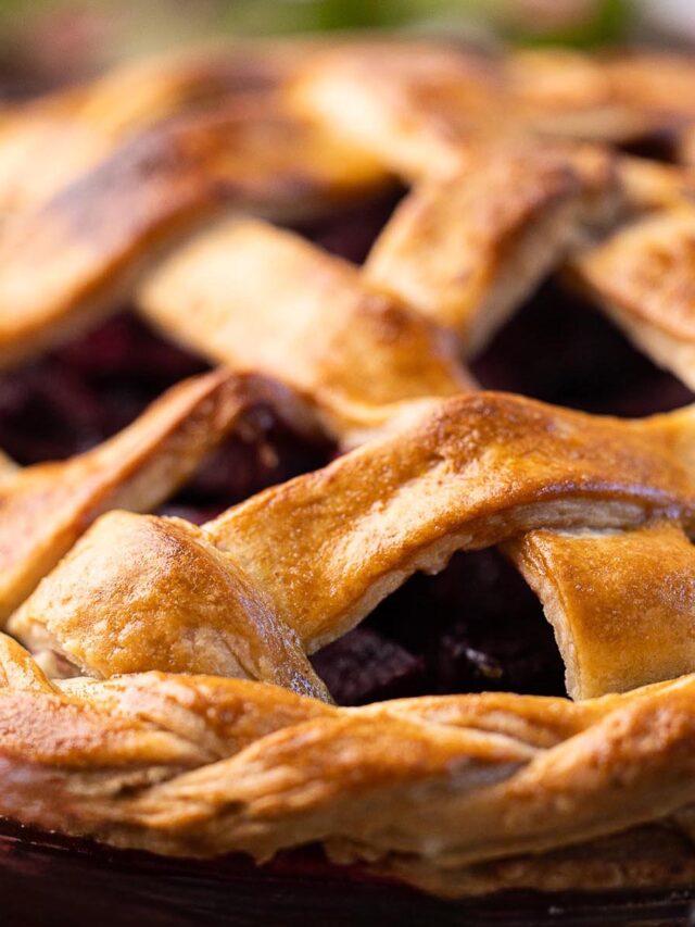 Golden, flaky lattice pie crust shown in close-up, revealing crisp texture and deep purple fruit filling beneath the woven top.