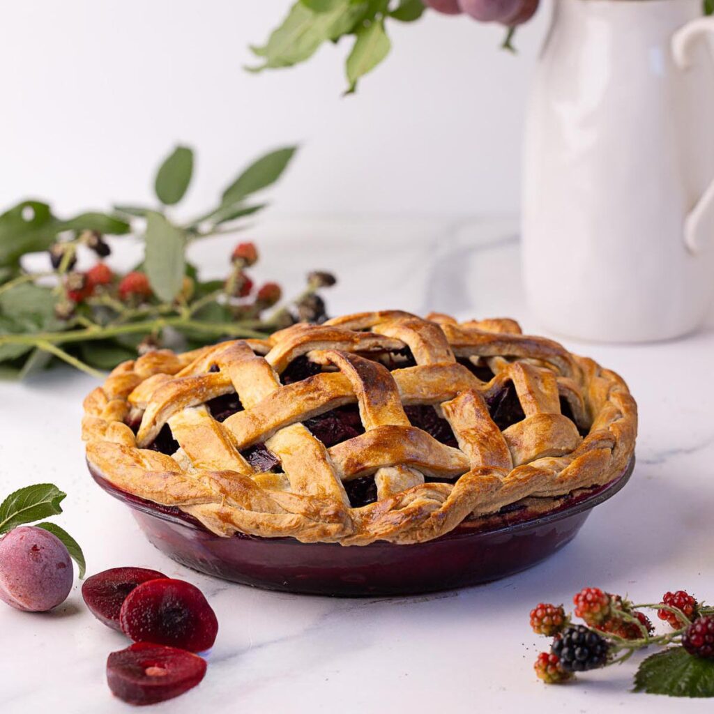 Blackberry plum pie with a golden lattice and braided crust, surrounded by fresh fruit and leaves on a marble surface, with a white pitcher in the background.