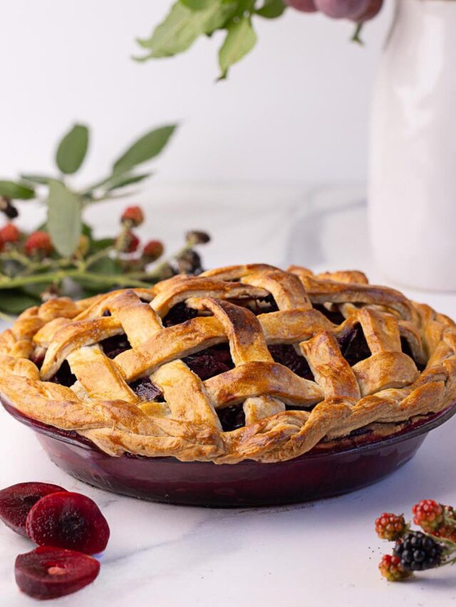 Blackberry plum pie with a golden lattice and braided crust, surrounded by fresh fruit and leaves on a marble surface, with a white pitcher in the background.