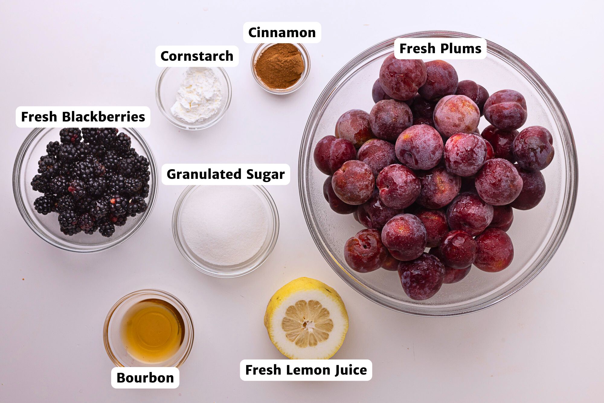 Labeled ingredients for blackberry plum pie filling displayed on a white surface, including fresh plums, blackberries, lemon juice, bourbon, granulated sugar, cinnamon, and cornstarch.
