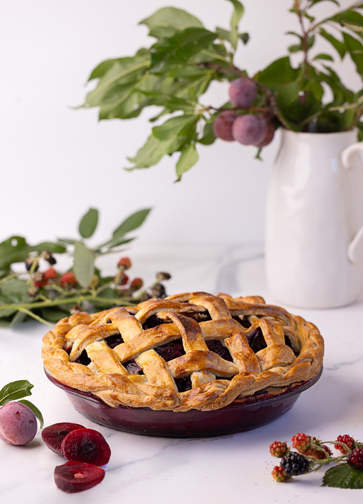 Fully baked blackberry plum pie with a golden lattice and braided crust, displayed on a white surface with fresh plums and blackberries, and leafy branches in the background for a rustic summer setting.