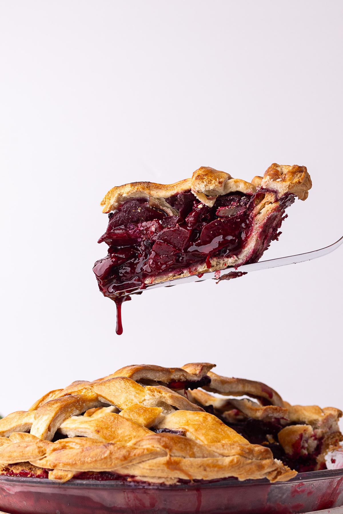 A slice of blackberry plum pie being lifted from the pie dish, with glossy filling dripping from the bottom and a golden lattice crust on top.