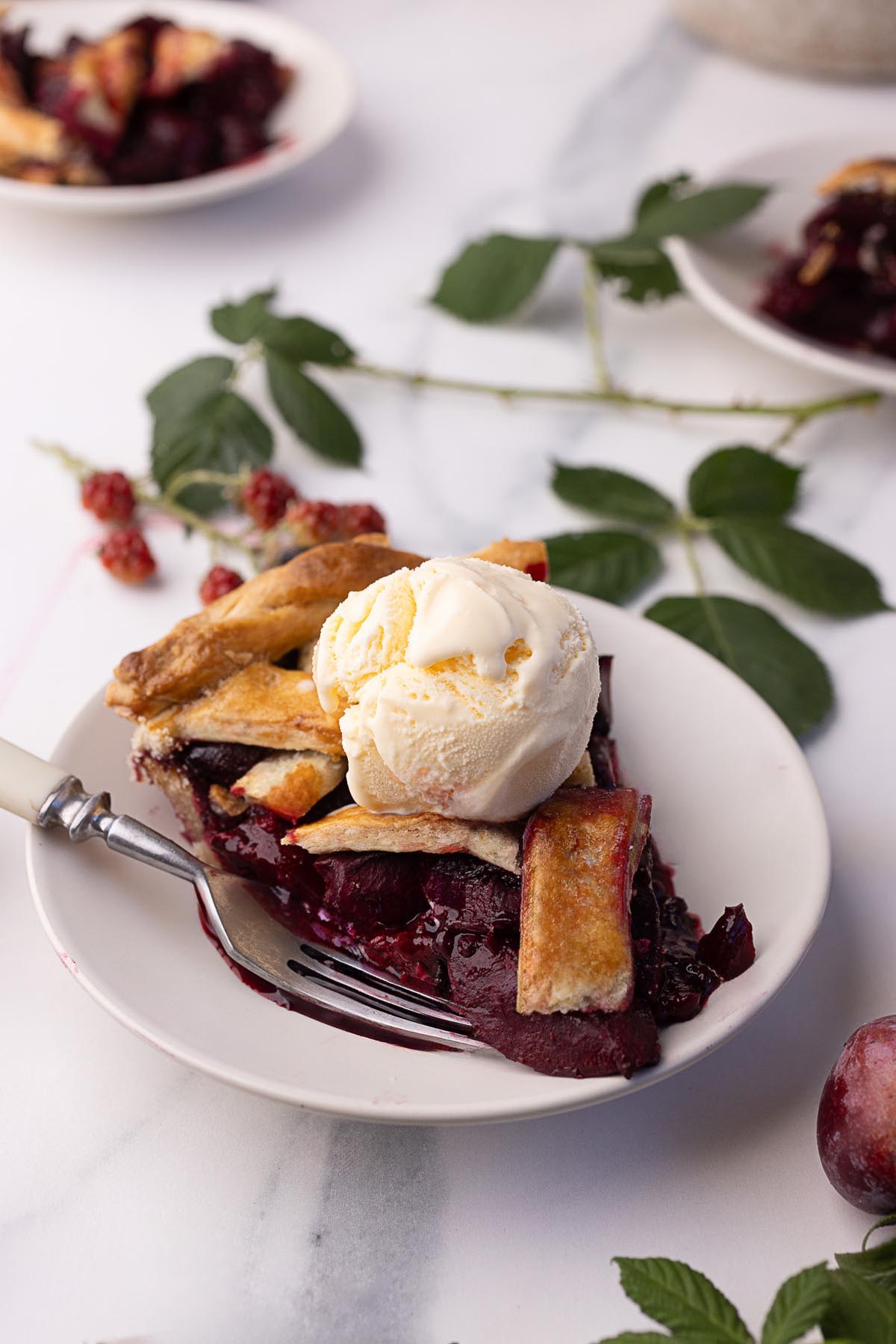Slice of blackberry plum pie topped with a scoop of vanilla ice cream on a white plate, with a fork and fresh blackberry leaves in the background.