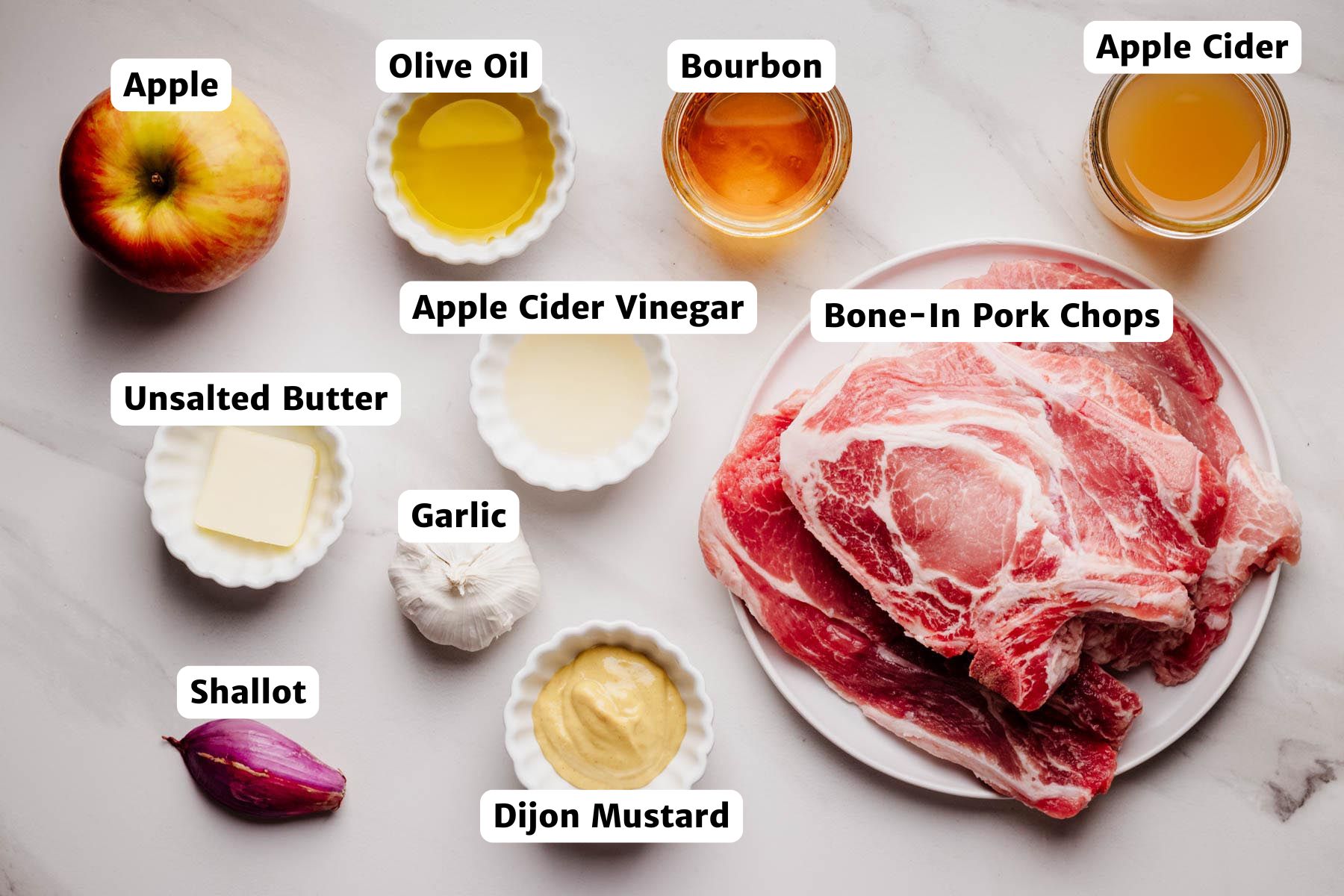 Flat lay of labeled ingredients for apple bourbon pork chops, including bone-in pork chops, apple, garlic, shallot, Dijon mustard, apple cider, apple cider vinegar, bourbon, olive oil, and unsalted butter.