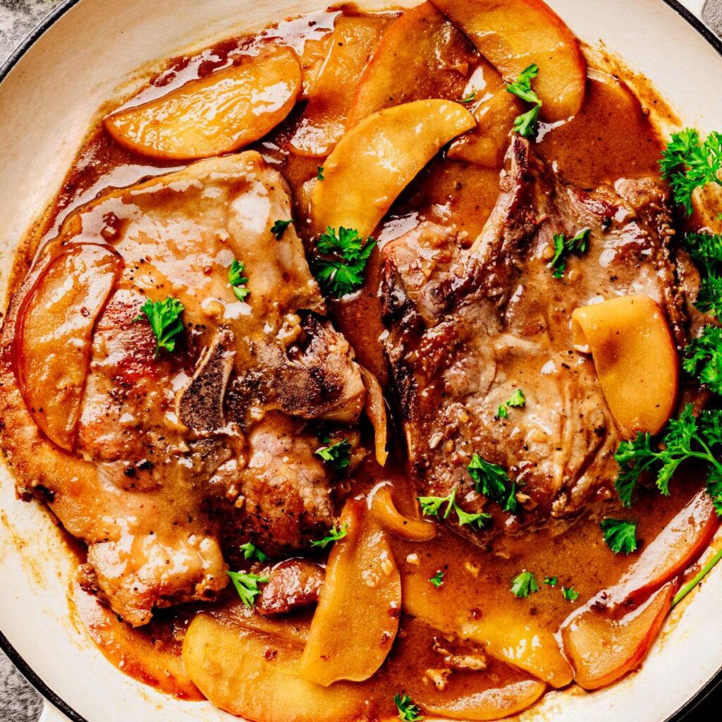 Two bone-in pork chops smothered in apple bourbon glaze with golden apple slices and fresh parsley in a white skillet, shot from overhead.
