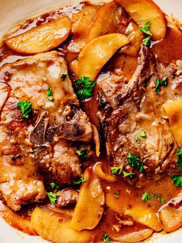 Two bone-in pork chops smothered in apple bourbon glaze with golden apple slices and fresh parsley in a white skillet, shot from overhead.