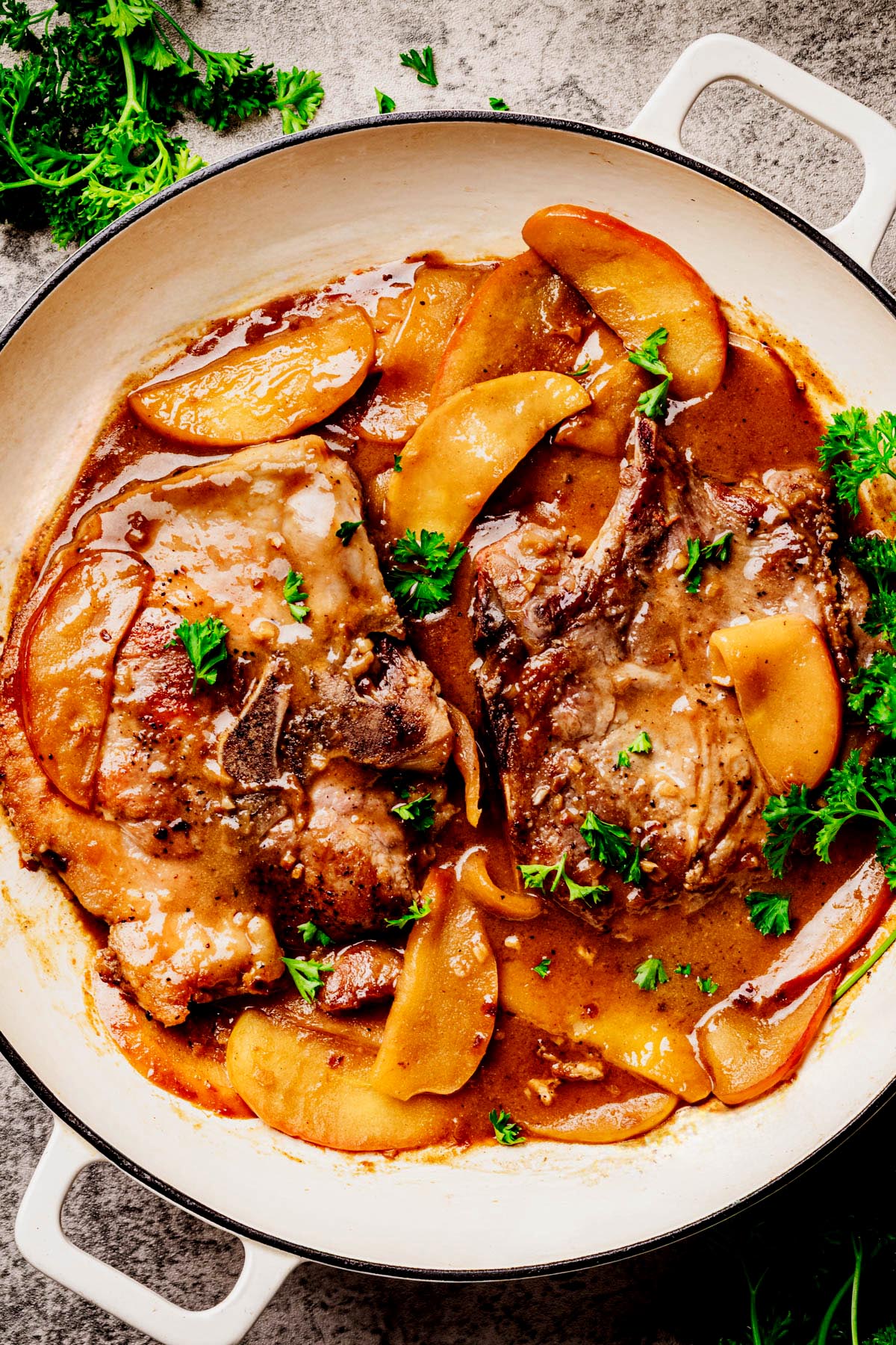 Two bone-in pork chops in a white skillet, simmered in a rich apple bourbon glaze with tender apple slices and garnished with fresh parsley on a stone surface.