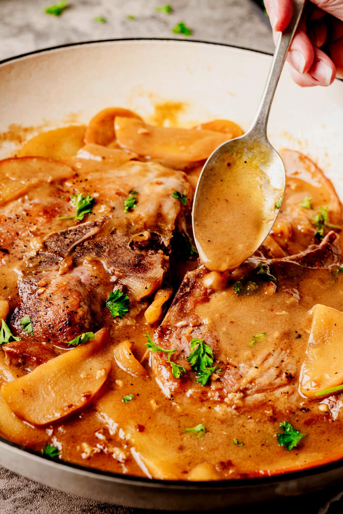 Close-up of bone-in pork chops in apple bourbon glaze, with a spoon drizzling sauce over the top, surrounded by tender apple slices and garnished with fresh parsley in a white skillet.