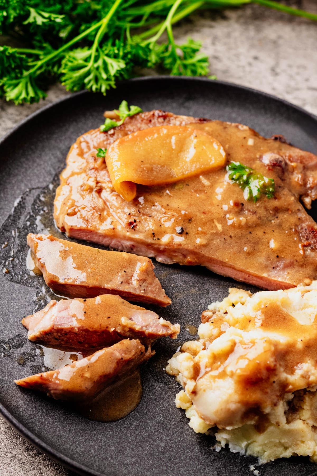 Sliced pork chop topped with apple bourbon glaze and apple slices, served with mashed potatoes and gravy on a dark plate, with fresh parsley in the background.