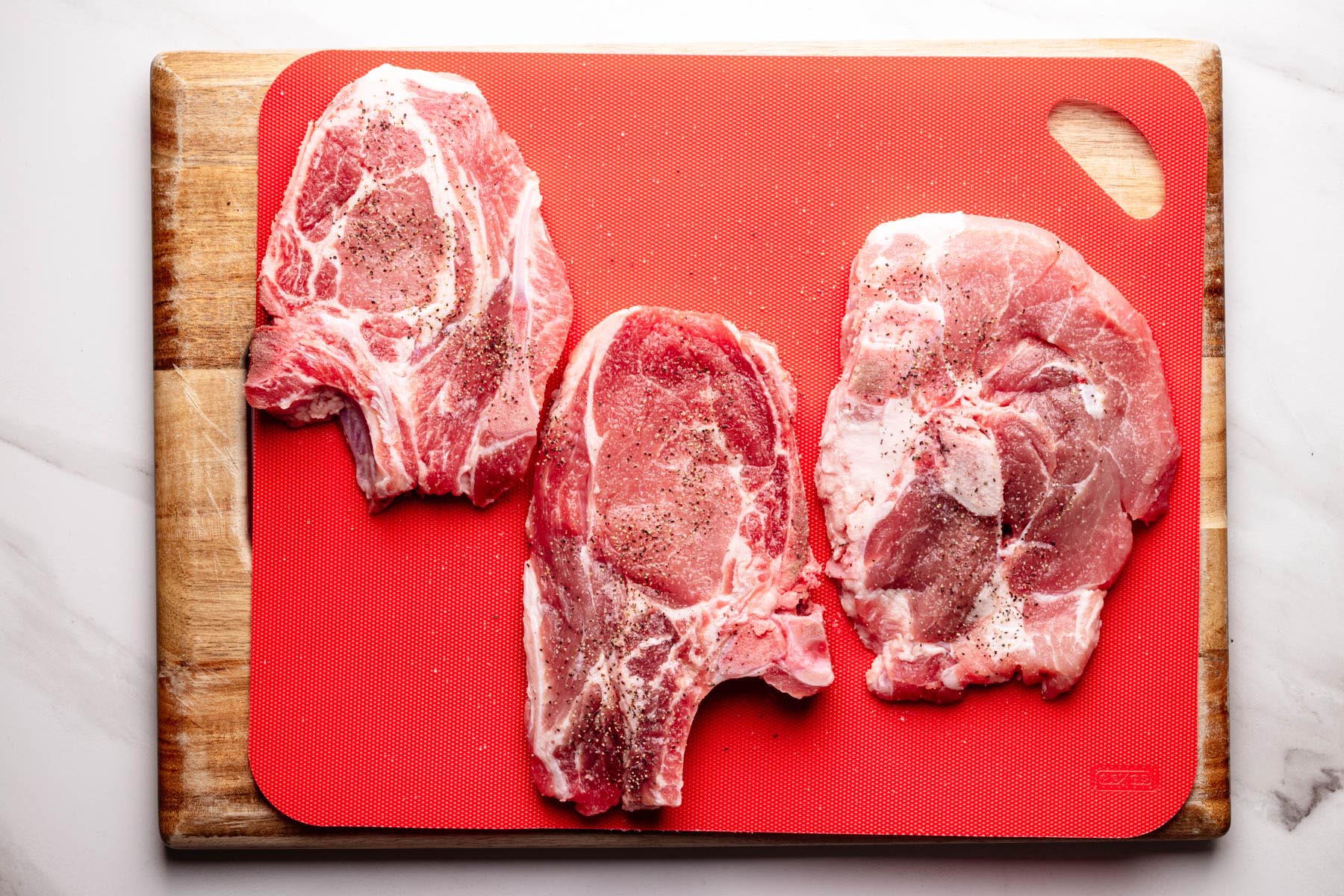 Three raw bone-in pork chops seasoned with salt and pepper on a red cutting board, placed on a wooden board over a white surface.