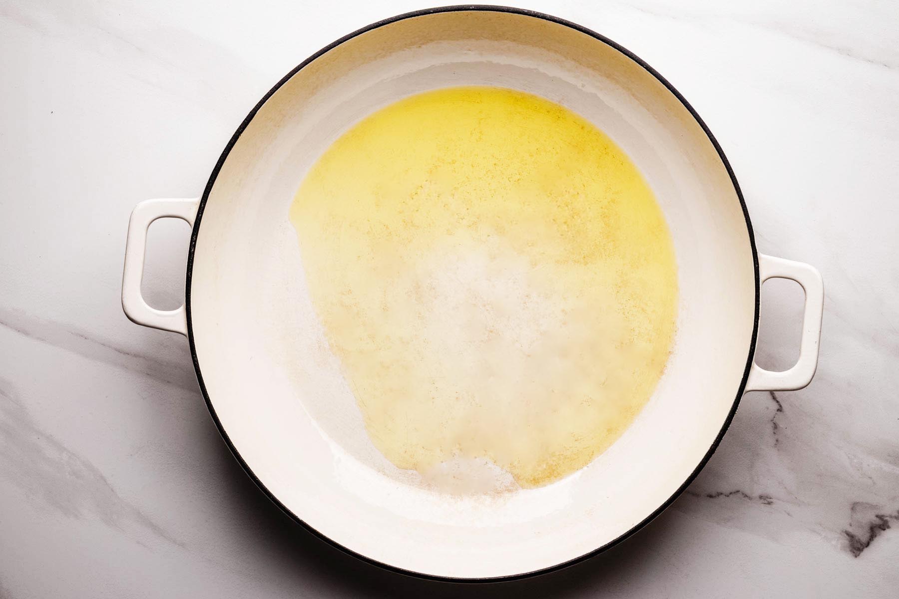 A white enamel skillet on a marble surface with hot olive oil shimmering in the center, ready for searing pork chops.