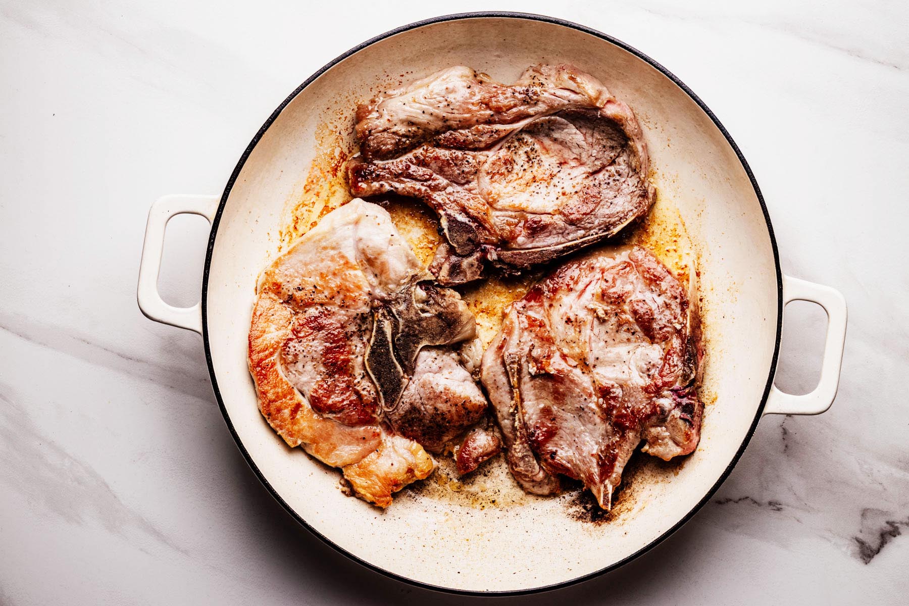 Three bone-in pork chops searing in a white enamel skillet, developing a golden-brown crust as they cook over medium-high heat.