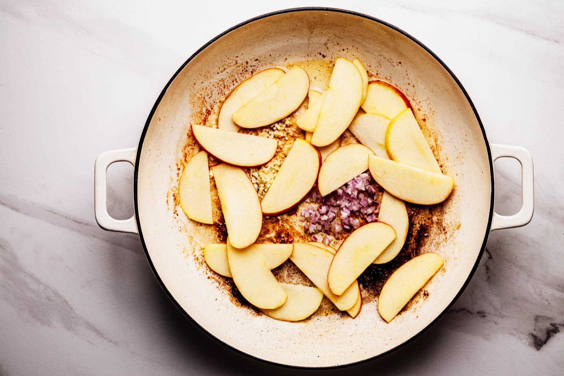 Thinly sliced apples, minced garlic, and chopped shallots sautéing in a white skillet, with browned bits from seared pork chops visible on the bottom of the pan.