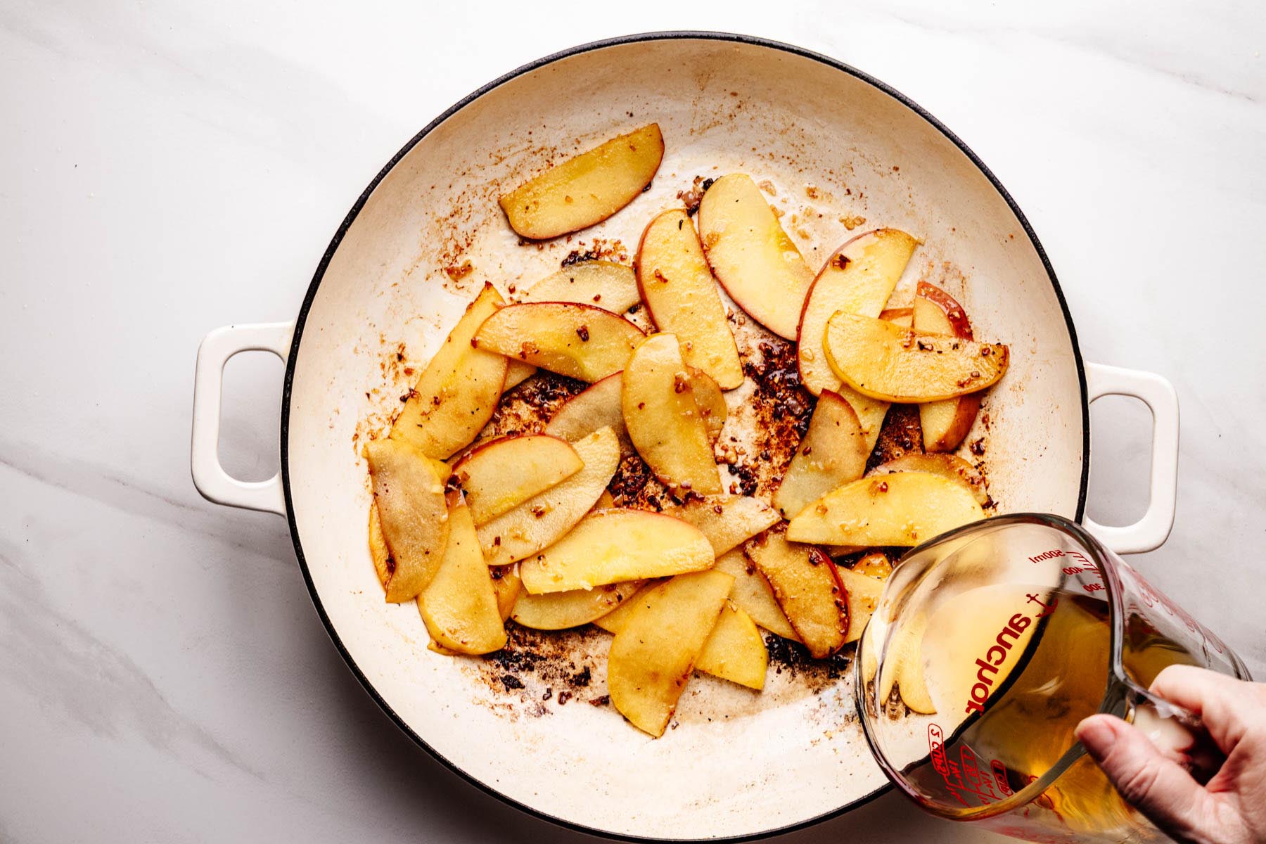 Sautéed apple slices in a white skillet with browned bits on the bottom, as a hand pours bourbon from a glass measuring cup into the pan to deglaze.