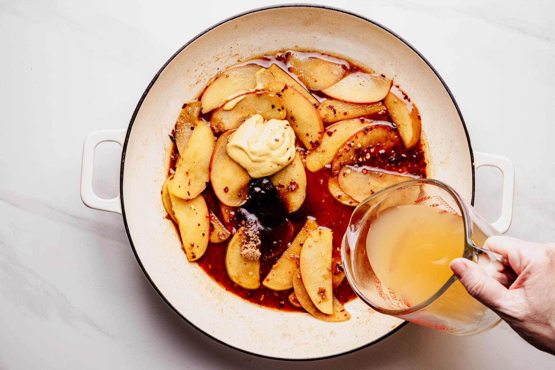A white skillet filled with sautéed apples and glaze ingredients, including Dijon mustard, brown sugar, and red pepper flakes, as apple cider is poured in from a glass measuring cup.