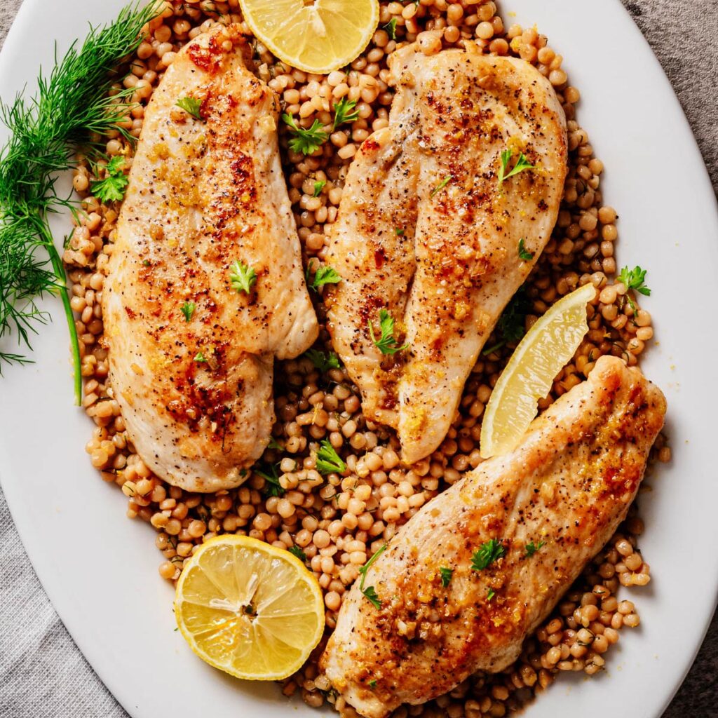 Overhead view of golden garlic butter chicken cutlets arranged on a bed of lemon dill pearl couscous, garnished with fresh herbs and lemon slices on a white serving platter.