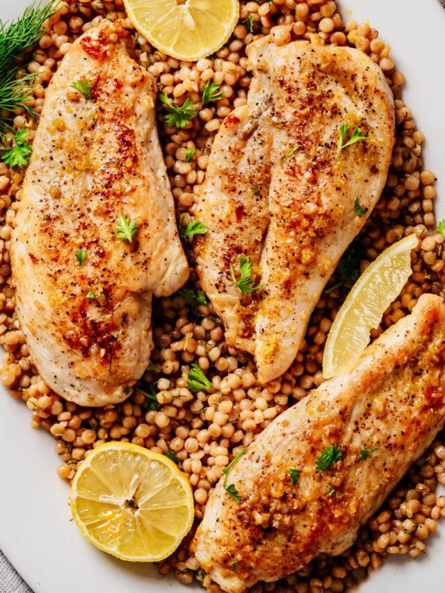 Overhead view of golden garlic butter chicken cutlets arranged on a bed of lemon dill pearl couscous, garnished with fresh herbs and lemon slices on a white serving platter.