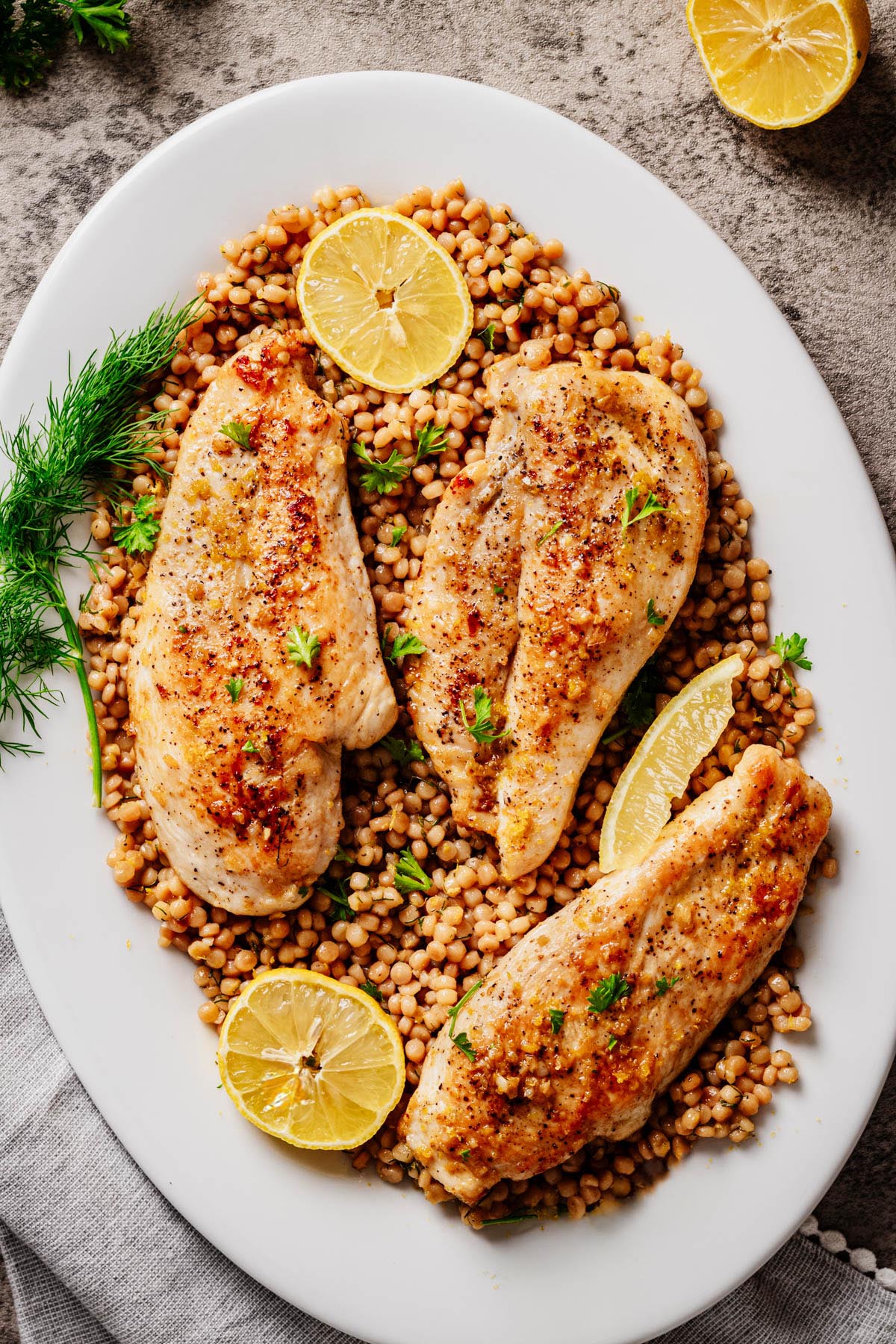 Three garlic butter chicken cutlets served over lemon dill pearl couscous on a white platter, garnished with fresh dill, parsley, and lemon slices.