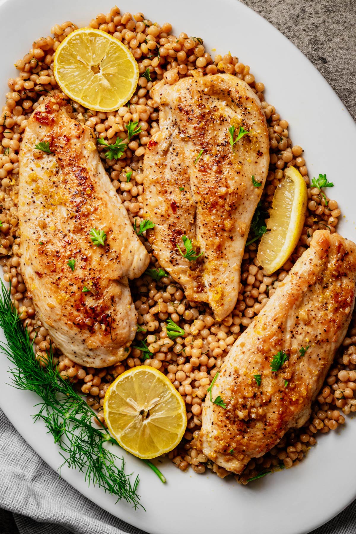 **Alt Text:** Close-up of garlic butter chicken cutlets on a bed of lemon dill pearl couscous, garnished with lemon slices, fresh dill, and parsley on a white platter.