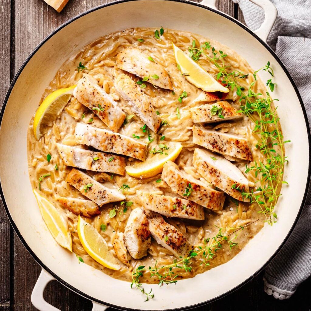 Creamy lemon chicken orzo pasta topped with sliced seared chicken breast, lemon wedges, and fresh thyme, served in a white braiser on a wooden surface.