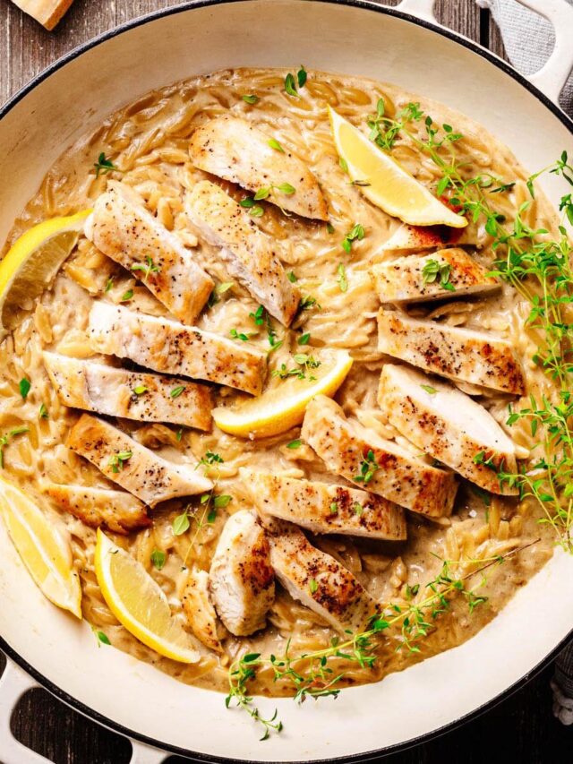 Creamy lemon chicken orzo pasta topped with sliced seared chicken breast, lemon wedges, and fresh thyme, served in a white braiser on a wooden surface.