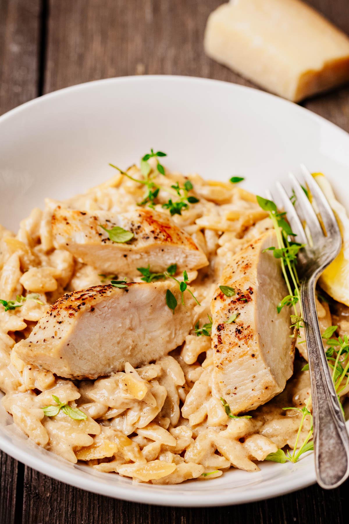 Creamy garlic-lemon orzo pasta served in a white bowl, topped with sliced seared chicken breast, fresh thyme, and a lemon wedge, with a fork resting on the side.