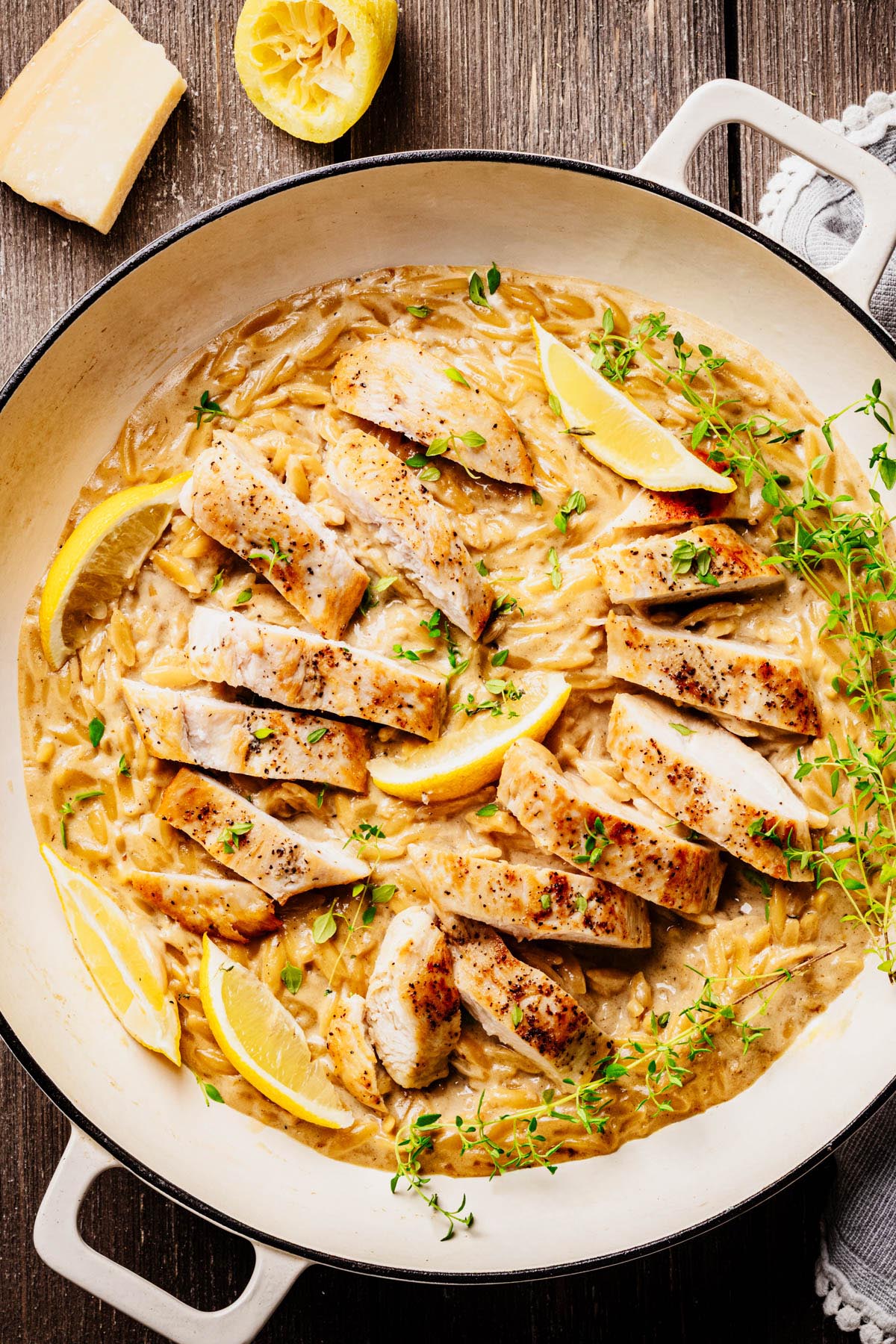 Creamy lemon chicken orzo pasta topped with sliced seared chicken, lemon wedges, and fresh thyme in a white braiser, with a wedge of Parmesan and squeezed lemon nearby.