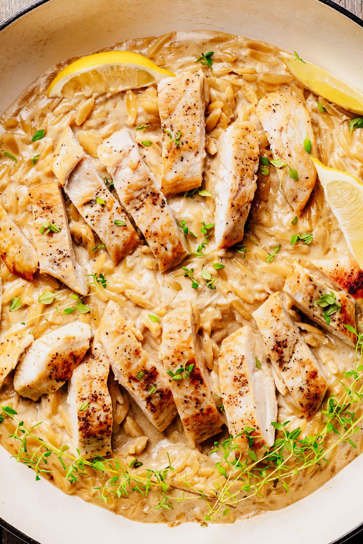 Close-up of creamy lemon chicken orzo pasta topped with sliced seared chicken, fresh thyme, and lemon wedges in a white braiser.