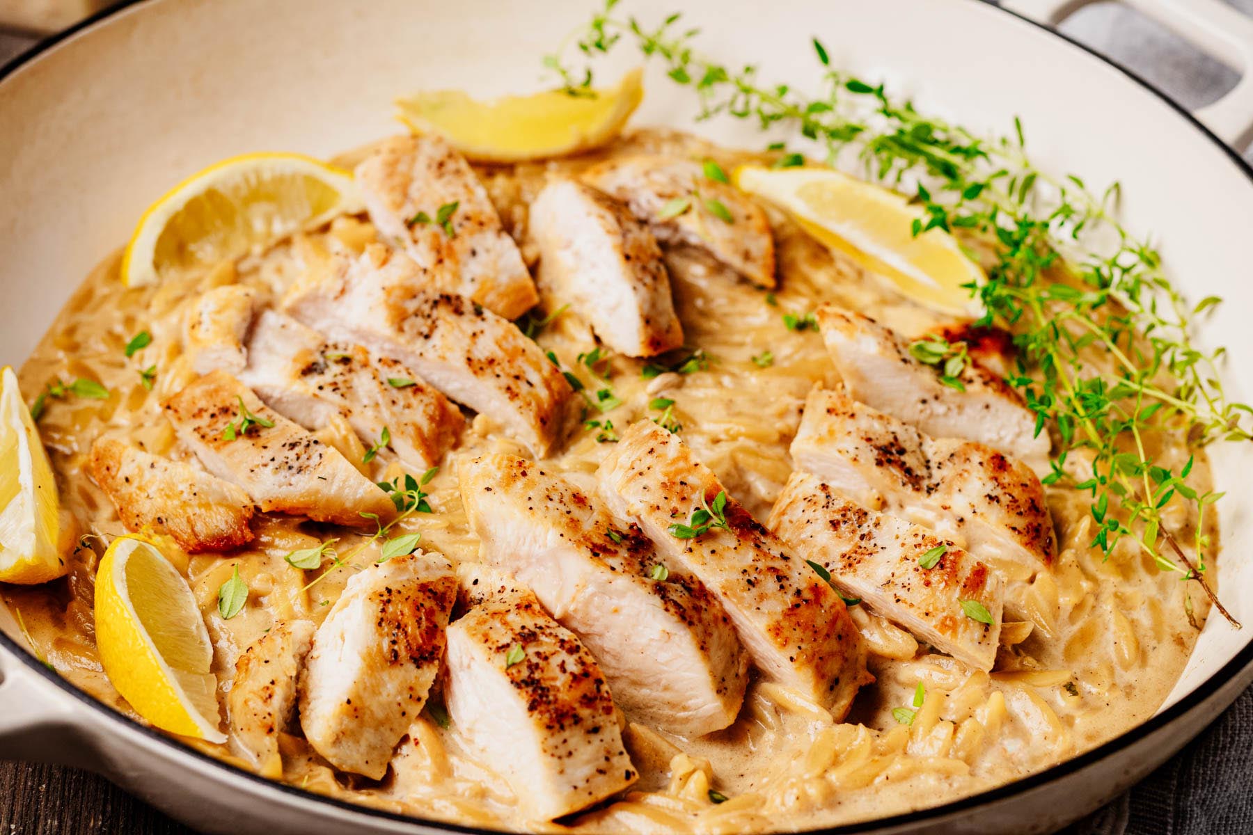 Creamy lemon chicken orzo pasta in a white braiser, topped with sliced golden-seared chicken breast, fresh thyme, and lemon wedges.