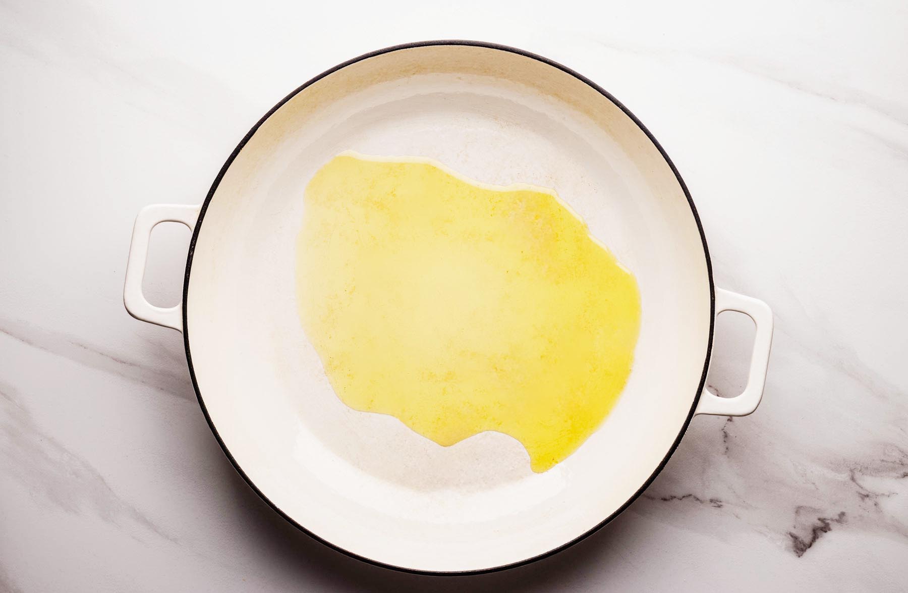 Olive oil heating in a large white braiser pan on a light marble surface, ready for cooking.