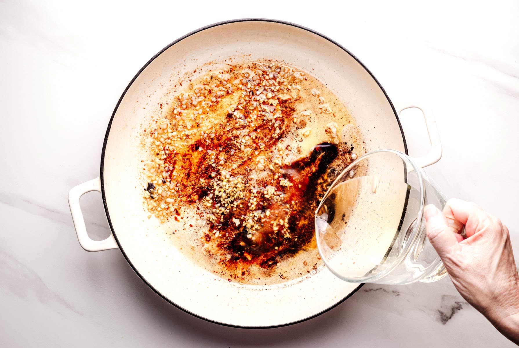 A hand pouring white wine into a white braiser pan with sautéed garlic, shallots, and browned bits from seared chicken to deglaze the pan.
