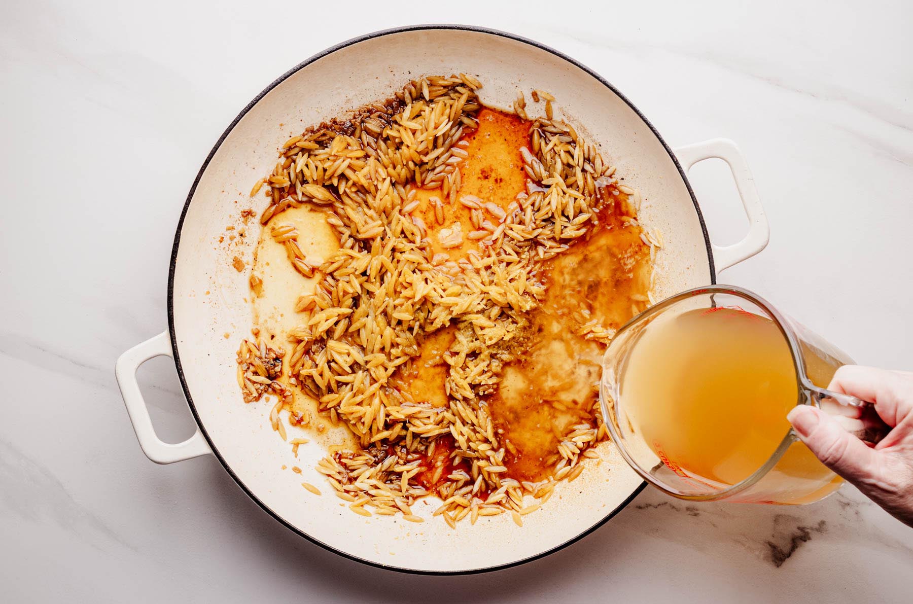 A hand pouring chicken broth into a white braiser pan containing toasted orzo and browned aromatics, preparing to simmer the mixture.