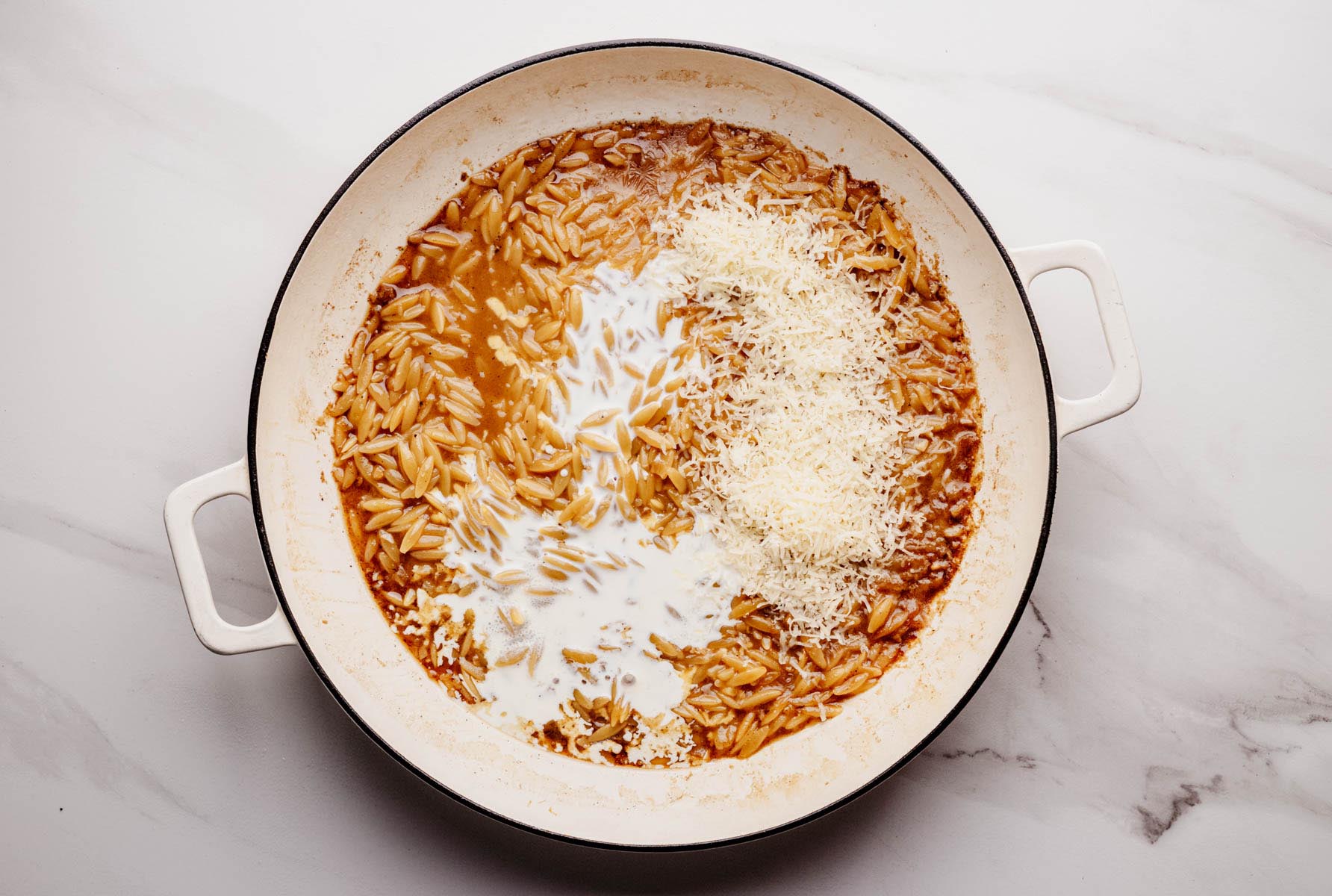 Simmered orzo pasta in a white braiser pan with added cream and freshly grated Parmesan cheese, ready to be stirred into a creamy sauce.
